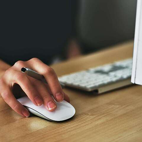 Eine Nahaufnahme der Hand einer Person, die eine weiße Computermaus benutzt. Die Hand liegt auf einem Holzschreibtisch in der Nähe einer Tastatur und eines Computermonitors.
