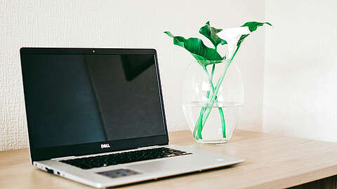 Ein geschlossener Dell-Laptop steht auf einem Holzschreibtisch neben einer durchsichtigen Glasvase mit grünen Blättern und weißen Calla-Lilien. Der Hintergrund ist eine helle, strukturierte Wand.