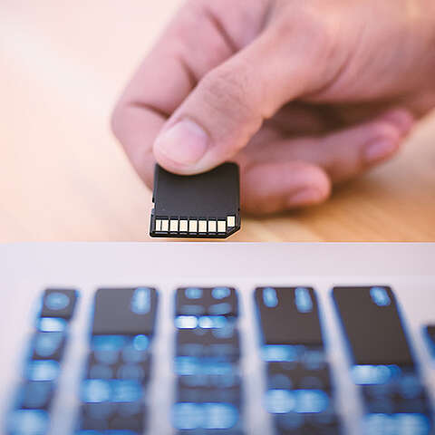 Eine SanDisk Extreme Portable SSD liegt neben ihrer geöffneten Verpackung auf einem Holztisch. Die SSD besticht durch ihr elegantes schwarzes Design mit einer orangefarbenen Schleife. Andere technische Geräte sind im Hintergrund unscharf dargestellt.