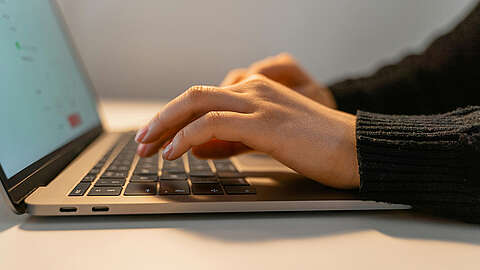 Eine Nahaufnahme der Hände einer Person, die auf einer Laptoptastatur tippt, wobei der Bildschirm leicht sichtbar ist und ein weicher, neutraler Hintergrund zu sehen ist. Die Person trägt einen dunklen, langärmeligen Pullover.