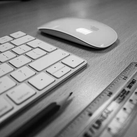 Ein Graustufenbild eines Schreibtischaufbaus mit einer kabellosen Tastatur, einer eleganten Maus, einem Holzstift und einem transparenten Lineal mit Zollangaben. Sie sind auf einer hellen Holzoberfläche angeordnet.