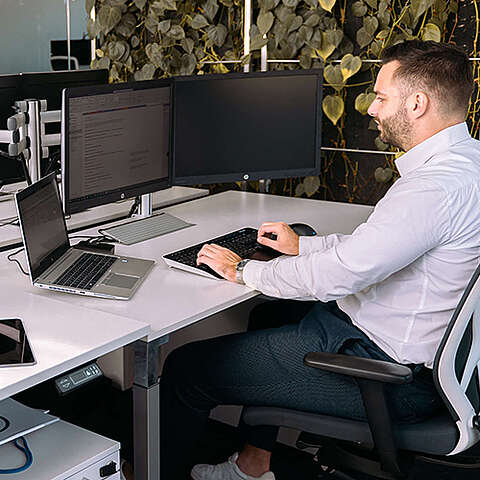 Ein modernes Home-Office mit einem Laptop auf einem Holzschreibtisch, einer eleganten Lampe und einer Pflanze in der Ecke. Eine French Press und eine Tasse stehen auf einem weiteren Tisch im Vordergrund, Licht fällt durch ein Fenster.