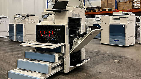 Ein großer weiß-blauer Bürodrucker mit geöffneten Schubladen, Seitenwänden und Fächern steht in einem Lagerhaus, umgeben von anderen ähnlichen Druckern und Regalen mit Kisten.