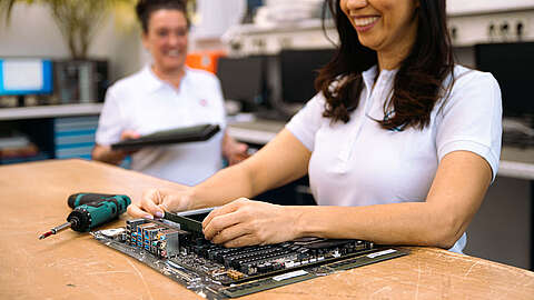 Zwei Frauen in weißen Hemden arbeiten in einem technischen Labor; eine baut lächelnd Computerhardware auf einem Tisch mit Werkzeugen zusammen, während die andere im Hintergrund einen Laptop hält.